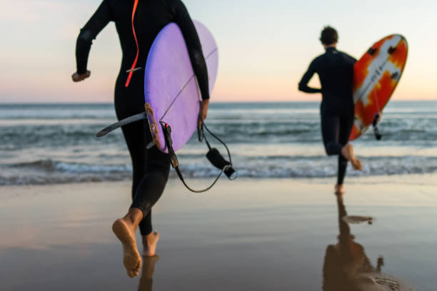 A man and a woman, clad in wetsuits, hasten along a sandy shore, surfboards in hand, under the fading light of a retiring sun.