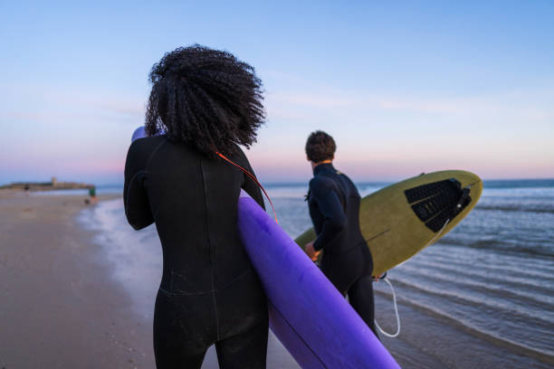 A diverse couple in wetsuits clutch their surfboards, energetically racing down a sandy beach as the sunset paints the sky with soft hues.