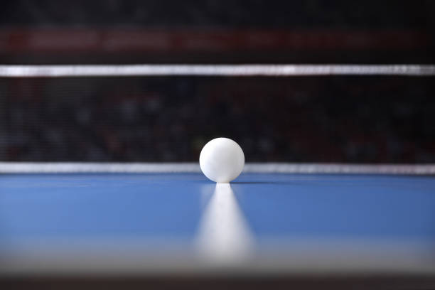 Ping pong competition concept with ball on the center line of a blue professional table with net and sports hall in the background. Front view.