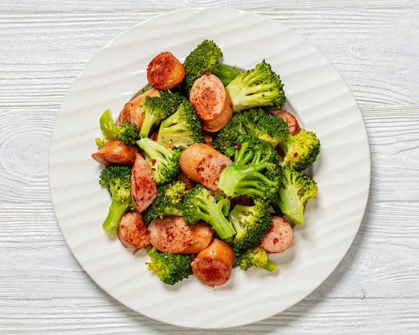 pan seared juicy sausage and crispy tender broccoli on white plate on white wooden table, horizontal view from above, flat lay, close-up