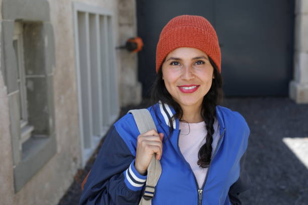 A very beautiful young female student in her twenties is walking on the street with her backpack. She wears an orange beanie and a classic university jacket.