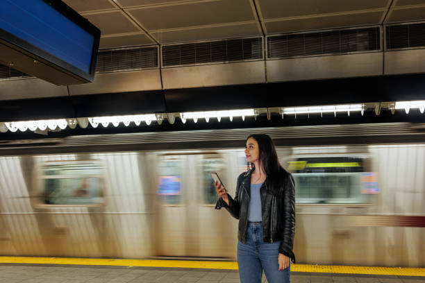 Young woman standing on the subway platform holding a phone while a train in blurred motion passes behind her.