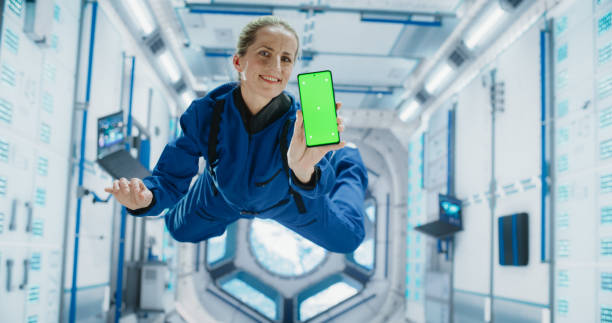 istock Female Astronaut Floating Inside Space Station in Zero Gravity. Spacewoman Showing a Smartphone with Green Screen Mock Up Template. Woman Posing for Camera, Smiling in Room in a Spacecraft 2156520031