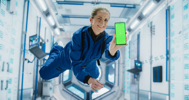 istock Female Astronaut Floating Inside Space Station in Zero Gravity. Spacewoman Showing a Smartphone with Green Screen Mock Up Template. Woman Posing for Camera, Smiling in a Room in a Spacecraft 2156520067