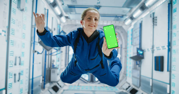 istock Female Astronaut Floating Inside a Space Station in Zero Gravity. Spacewoman Showing a Smartphone with Green Screen Mock Up Template. Woman Posing for Camera, Smiling in a Room in a Spacecraft 2156520096