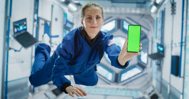 istock Caucasian Female Astronaut Floating Inside Space Station in Zero Gravity. Spacewoman Showing a Smartphone with Green Screen Mock-Up Template. Woman Posing for Camera, Smiling in Room in a Spacecraft 2156520464