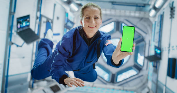 istock Caucasian Female Astronaut Floating Inside Space Station in Zero Gravity. Spacewoman Showing a Smartphone with Green Screen Mock Up Template. Woman Posing for Camera, Smiling in Room in a Spacecraft 2156520492