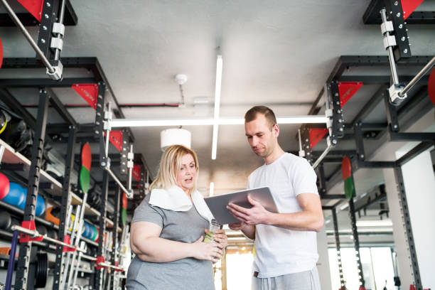 istock Overweight woman consulting with trainer, discussing daily workout plan. Personal trainer talking with client, discussing exercise program in gym. 2156546933