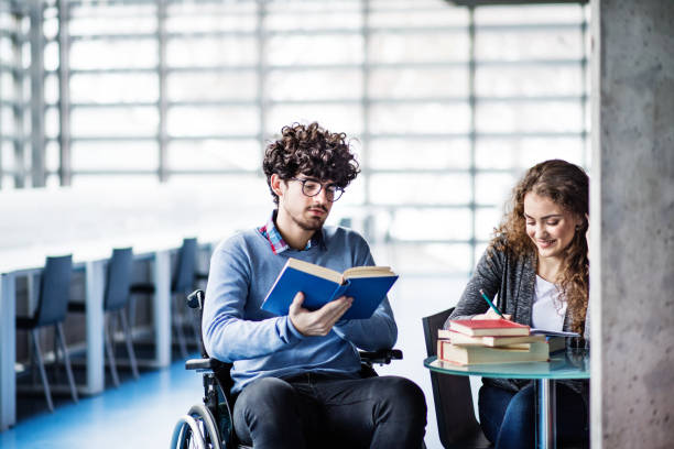 istock Handsome university student in wheelchair studying in library with classmate, preparing for final exam, presentation. Study group. Teamwork skills. 2156552632