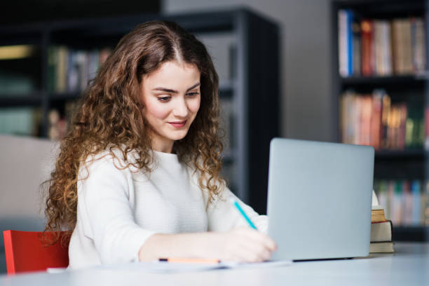 istock Young female student in library, focusing on final project, presentation, working on laptop. University student preparing for final exam. 2156553223
