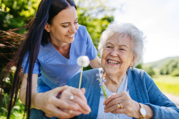 Female caregiver and senior woman in wheelchair holding dandelion, picking wild flowers. Nurse and elderly woman enjoying a warm day in nursing home, public park. Caregiver and senior woman in wheelchair holding dandelion, picking wild flowers. Nurse and elderly woman enjoying a warm day in nursing home, public park. nursing home stock pictures, royalty-free photos & images