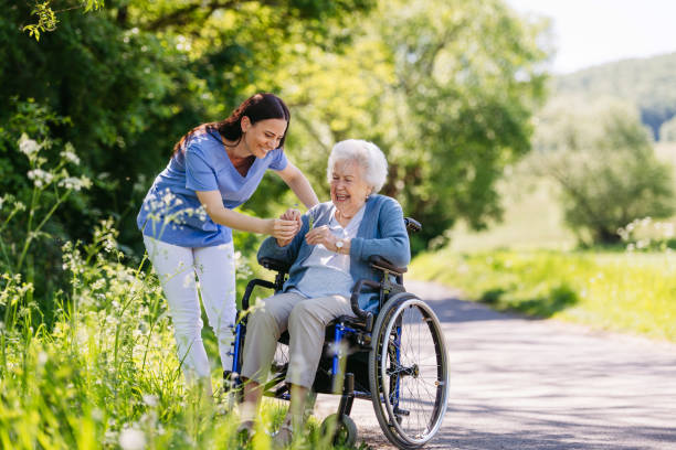 female caregiver and senior woman in wheelchair picking wild flowers. nurse and elderly woman enjoying a warm day in nursing home, public park. - heilberuf stock-fotos und bilder