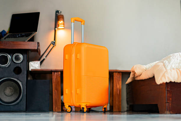 A bright yellow suitcase with a retractable handle stands on the floor next to a bed in a cozy room. The room features a wooden desk with a laptop and a lamp, and a speaker is visible on the floor.