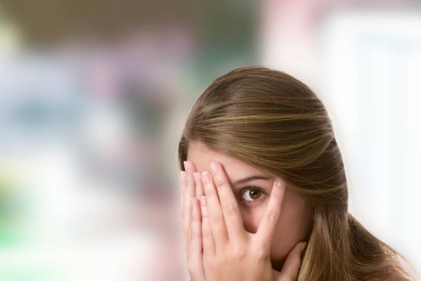 Facial expressions themes: Hispanic young woman with peeking through fingers expression and both hands on face.