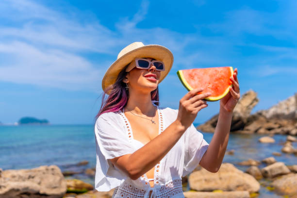 istock A woman is holding a watermelon slice in her hand while standing on a beach 2156610405