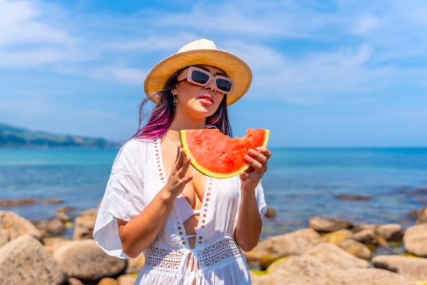 istock A woman is standing on a beach holding a watermelon slice 2156610421