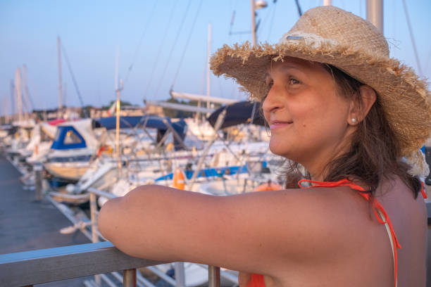Beautiful young woman in a marina. She is wearing a sleeveless orange dress and a yellow straw hat.