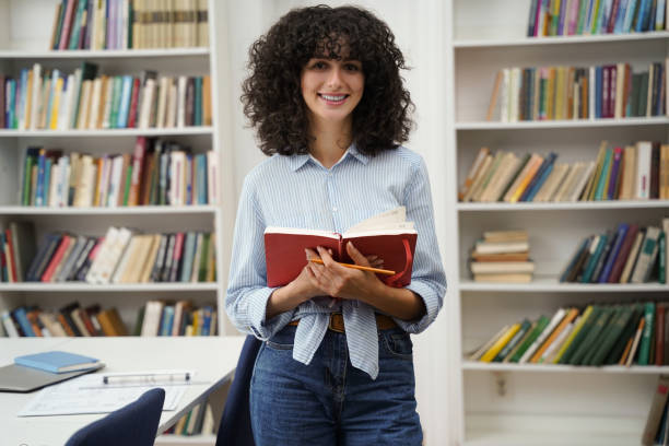 istock Curly-haired woman standing in the library with a book in hands 2156627267