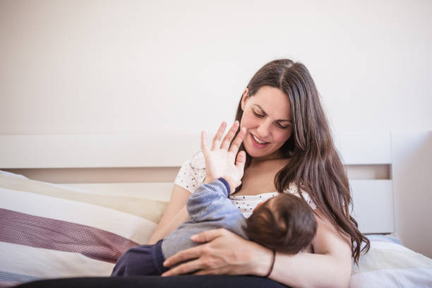 Loving mother breastfeeds her infant while sitting on a bed in the morning light, showcasing a tender moment of nurturing and bonding.