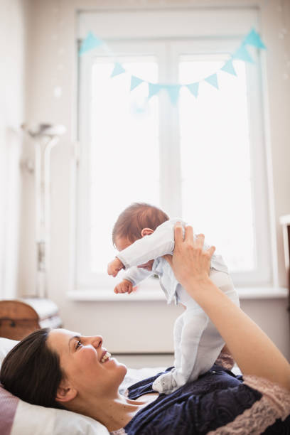 A joyful moment captured as a mother lifts her baby while lying down in a bright room with a window and blue bunting in the background, showcasing a serene and loving home environment.