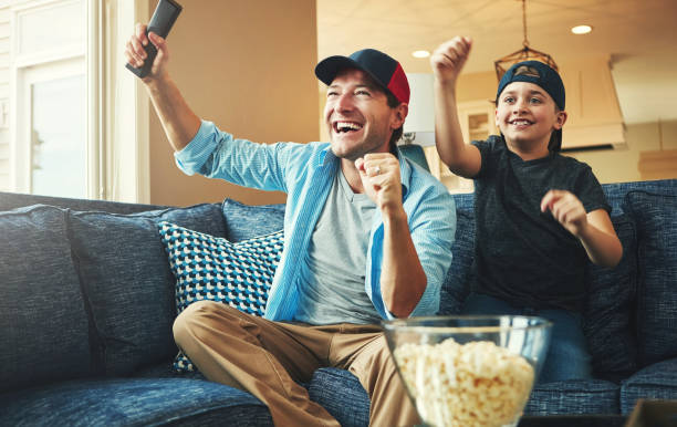 istock Television, kid and man with celebration on sofa for streaming, subscription and watching together. Son, dad and popcorn with tv remote in fist for bonding, success and live broadcast of sports match 2156718172