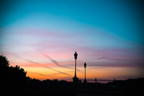 A beautiful sunset with vibrant colors of orange, pink, and blue in the sky. The silhouettes of vintage street lamps and trees are visible in the foreground, adding contrast and depth to the serene scene.