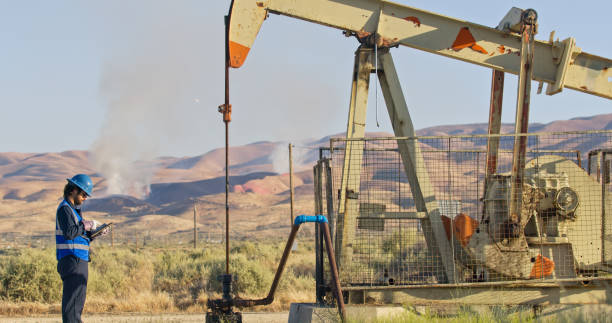 An Indian man in a blue hi-vis and helmet next to an oil pump jack on a sunny day in Kern County, California.