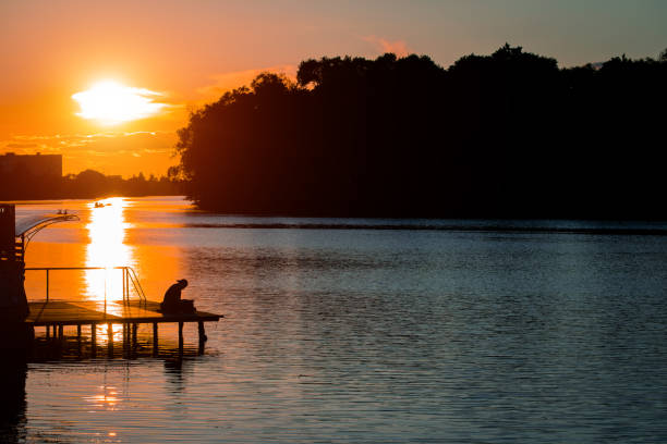 thinking alone unrecognizable in waterfront beach area panorama sunset landscape solitude atmosphere in darkness twilight lighting on river water surface from orange sky background, empty copy space