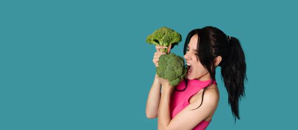 istock A slender smiling Caucasian young woman bites fresh broccoli on a blue background. Panorama, a place for text 2156743199