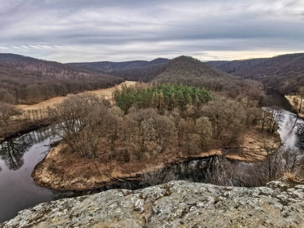 istock landscape from a bird's eye view. We see a meandering river that cuts through a valley with wooded hills. The prominent hill in the middle is densely covered with coniferous trees 2156754136