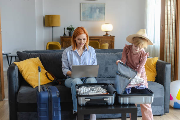 istock Getting ready for a summer holiday with child. Mother is using laptop while her daughter packing clothing in suitcase 2156758994