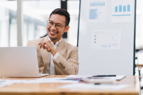 istock A handsome, smiling businessman wearing glasses and a suit, sitting at a desk while working on a laptop. 2156765976