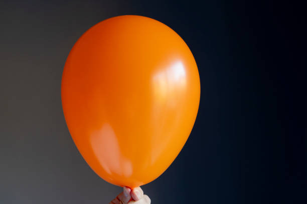Senior man holding up an orange balloon against a grey/gray wall.