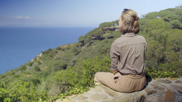 Young woman with brown hair stands by calm ocean under clear sky. Soft light highlights her thoughtful expression. Vivid colors enhance tranquil backdrop