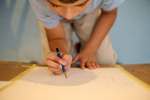 The hands of a child are focused on a drawing with a blue pencil, creating a robot image on paper that is partially finished with intricate lines
