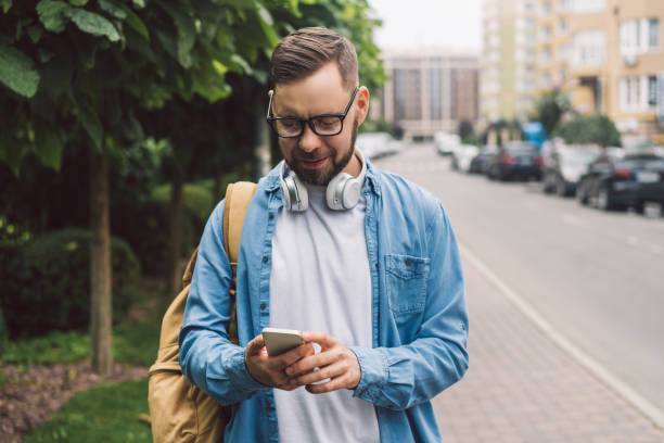 istock Portrait of a young smiling casually dressed handsome man sending message via his mobile phone while walking outdoors 2156814432