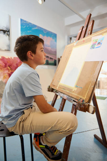 istock Young boy engaged in art class, drawing and painting on easel in a creative workshop 2156828613
