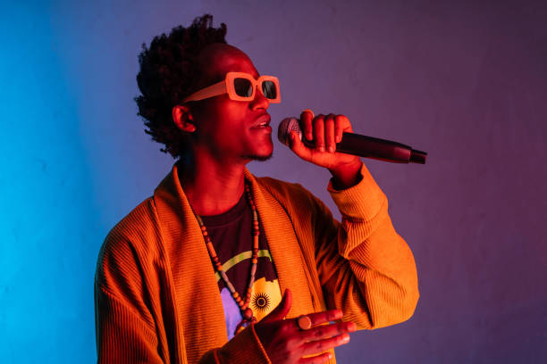 A smartly dressed African-American man is holding a microphone to his mouth as he sings against a blue to purple gradient background