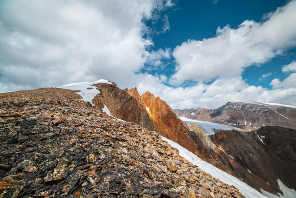 istock Snow-capped mountain ridge with cornice and red sharp rocks on top in sunlight. Snow cap and shiny pointy peak of gold color. Snow dome and colorful rocky peaked top. Sunny cloudy high mountains view. 2156838171