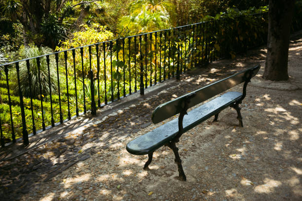A solitary green park bench in a shaded area near a wrought iron fence, surrounded by lush greenery and dappled sunlight. The ground is covered with small stones and dirt providing a serene atmosphere
