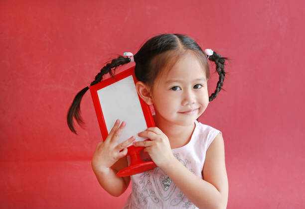 Adorable little asian girl holding empty whiteboard menu against red wall background.