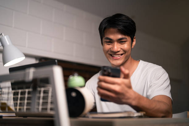 istock Asian young man overworking and using mobile phone check news at night. Attractive handsome businessman typing on smartphone while working to communicate online on table during bedtime in house. 2156863597