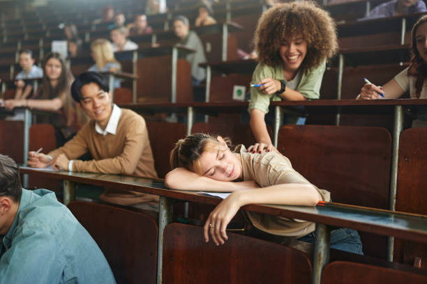 Come on, wake up, we don't sleep on a class! Happy female student trying to wake up her friend who fell asleep on a class at lecture hall. Focus is on woman sleeping. male college student sleeping through lecture stock pictures, royalty-free photos & images