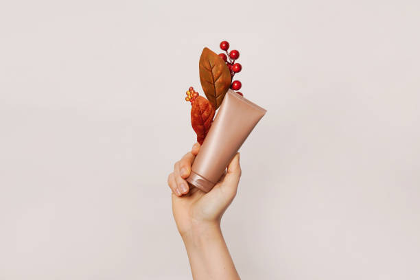 A hand holding a tan-colored cosmetic tube adorned with autumn-themed decorations including red berries and brown leaves