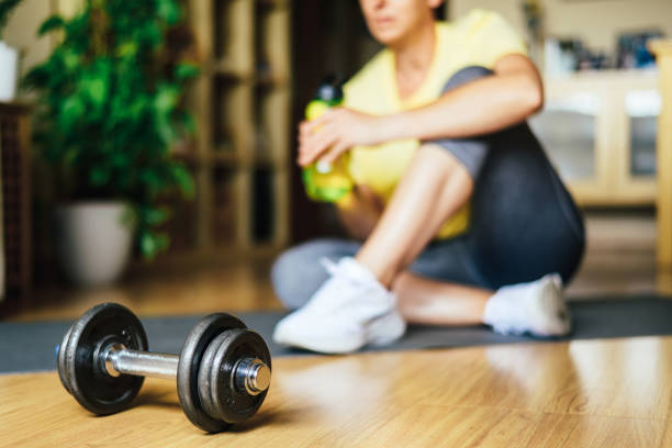 istock Person Taking A Break Drinking Water After Doing Her Daily Exercise At Home, Using Dumbbells And Mat, Wearing Sportswear In Yellow And Green Tones. Natural Light. Morning Routine And Staying Fit 2156874387