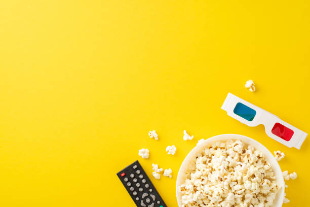 istock Home theater vibes: top view of popcorn, 3D glasses, and remote control for streaming. Ideal for promotions or personalized messages against bright yellow 2156875202