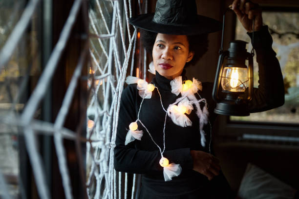 Pensive African American woman dressed up as a witch on Halloween looking through window from home.