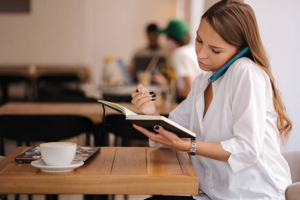 Financial manager writing down in notebook and speak by phone in same time. Working in cafe. Freelancer concept. High quality photo