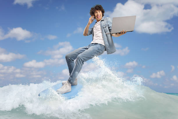 Young man in jeans on a surfboard using a smartphone and holding a laptop computer in the sea