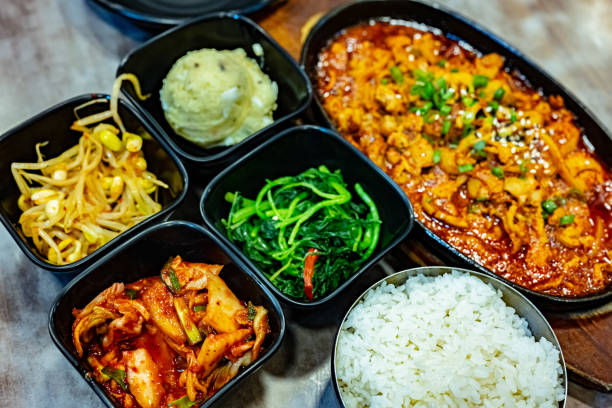 A close-up shot of a person's hands using chopsticks to place a piece of fresh fermented kimchi onto a fresh green leaf. Out-of-focus side dishes and sauces, highlighting the authentic and communal experience of Korean barbecue.